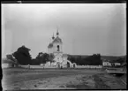 'Vy med kyrka på avstånd, samma kyrka som fotonr. 5282:10. Människor på öppen plan framför kyrkan. Till höger liten bro över uttorkat vattendrag / dike, 1 man stående på bron. ::  :: Ingår i serie med fotonr. 5282:1-15. Se även fotonr. 5269-5292 med bilder från Stuxbergs resa till Kaukasien.'