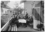 'Enligt anteckning på baksidan av fotografiet: ''Orvar Nybelin halvsittande bakerst, i mitten på Kristineberg, ca 1913-1915.'' ::  :: 10 st män sittande kring ett bord på en veranda, iklädda kostym och studentmössa.'