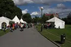 'Göteborgs Naturhistoriska Museums deltagande i  ''Skogens och Jordens dag'' i Slottsskogen. Museets tält uppsatt nära Linnéplatsen. ::  :: Vy mot Linnéplatsen från Slottsskogen med vita tält uppsatta på båda sidor om gång- och cykleväg, på gräsmatta. Människor gående på vägen. Främst till höger är museéts tält. Papperskorg, gatubelysning. ::  :: Ingår i serie med fotonr. 7074:1-4.'