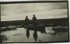 'Tobi Tourunen och forstmästare Henry Hackman stående i roddbåt. Vy över sjö med vegetation, vass. I fonden syns hus. ::  :: Serie fotonr 953-971.'