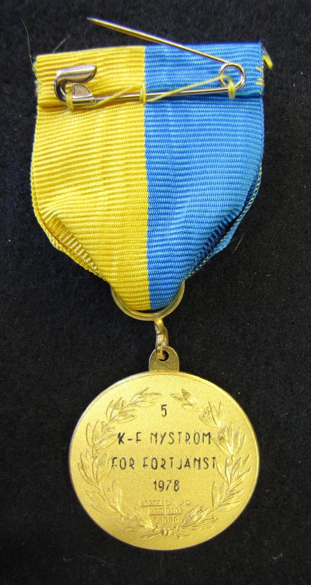 Medalj med sidenband. Västergöta-Dals pistolskyttekrets medalj tilldelad Karl-Fredrik Nyström. 5 K-F Nyström för förtjänst 1978.

På åtsidan finns två korslagda pistoler. Bakom dem finns en lagerkrans. Ovanför två sköldar, en med Västergötlands vapen och den andra en med Dalslands vapen. Runt kanten står Västgöta-Dals pistolskyttekrets.

Medaljen ingår i en samling märken och medaljer sittande på en tavla med märken, framförallt skyttemärken.

Karl-Fredrik Nyström var aktiv fritidsskytt och var en av de drivande för att starta Vänersborgs skytteklubb.