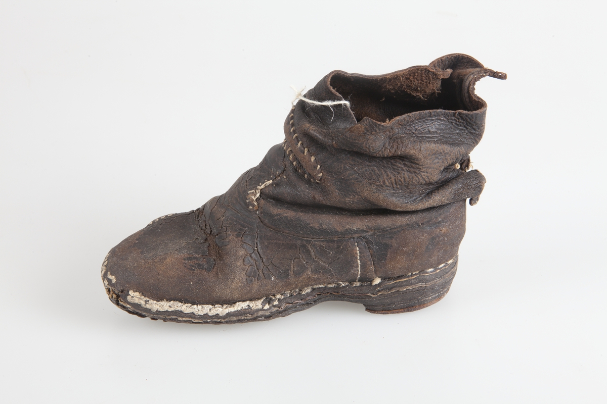 Udda barnkänga. Enläst. Brunsvart läder, nävermellansula. Handsydd. Randsydd, reseft, söm bak. Träpliggad sula och klack, klackjärn. Skaftet avskuret. Trasig, lagad.