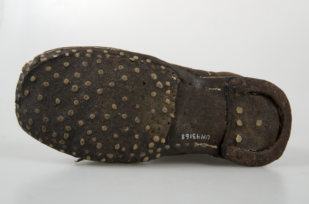 Udda barnkänga. Enläst. Brunsvart läder, nävermellansula. Handsydd. Randsydd, reseft, söm bak. Träpliggad sula och klack, klackjärn. Skaftet avskuret. Trasig, lagad.