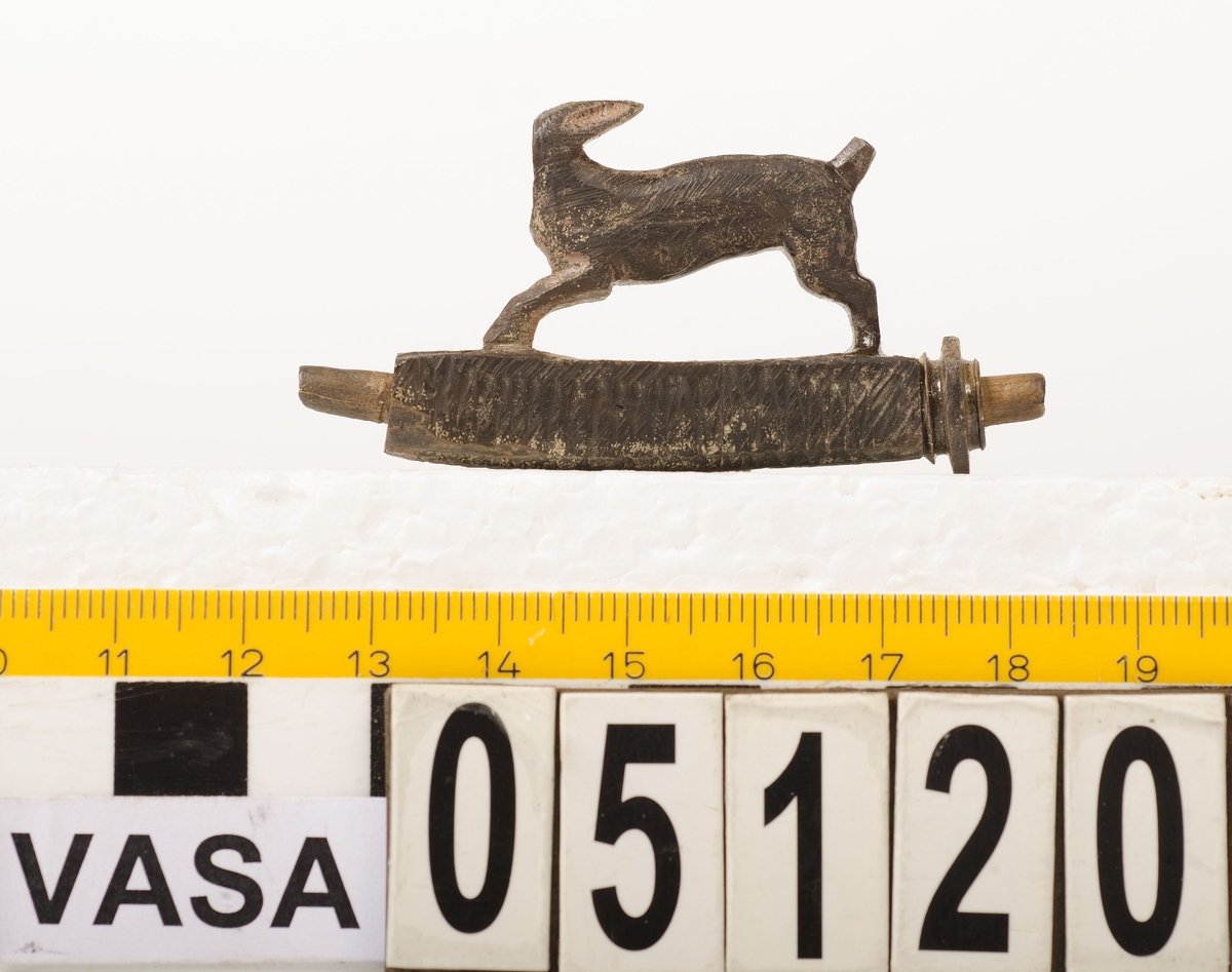 Snidad djurfigur. Basen som djuret står på har kvadratiskt tvärsnitt. Genom basen löper ett ihåligt rör. Djurfiguren föreställer en löpande hare, hjort eller hund. Djurhuvudet är avbrutet.