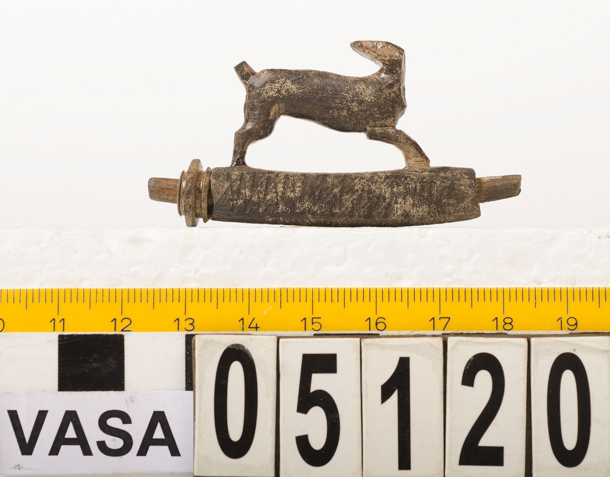 Snidad djurfigur. Basen som djuret står på har kvadratiskt tvärsnitt. Genom basen löper ett ihåligt rör. Djurfiguren föreställer en löpande hare, hjort eller hund. Djurhuvudet är avbrutet.