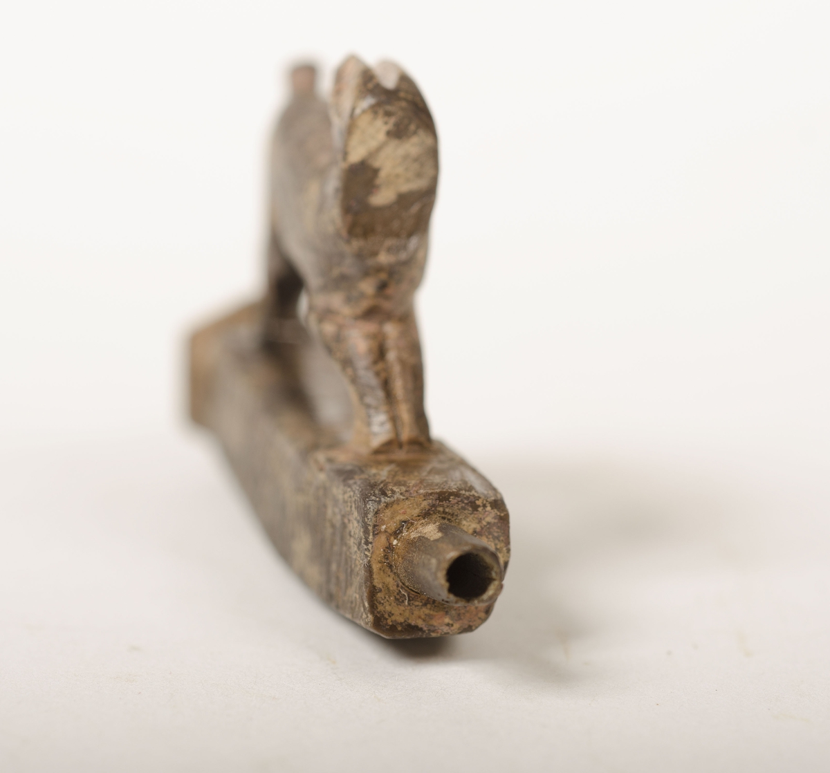 Snidad djurfigur. Basen som djuret står på har kvadratiskt tvärsnitt. Genom basen löper ett ihåligt rör. Djurfiguren föreställer en löpande hare, hjort eller hund. Djurhuvudet är avbrutet.