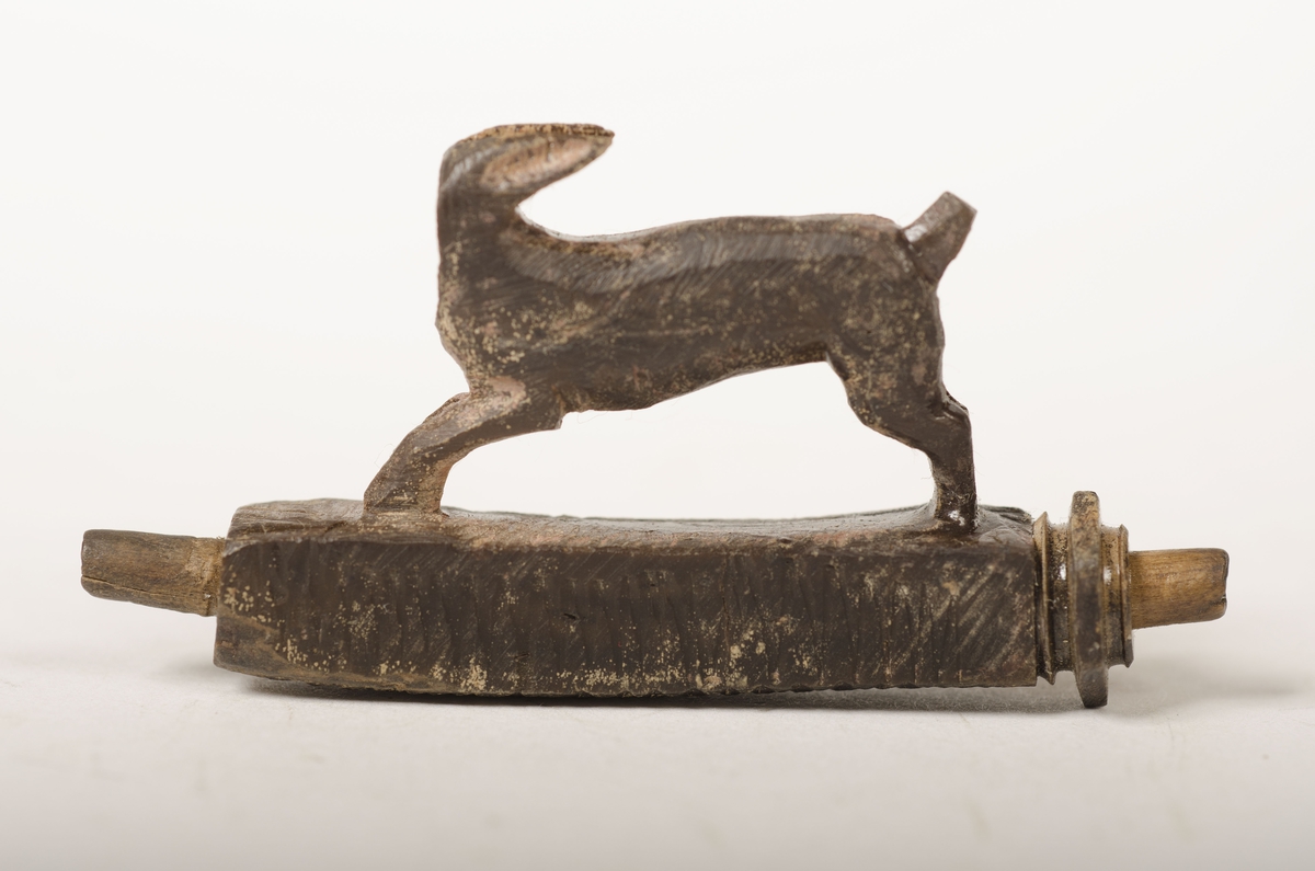 Snidad djurfigur. Basen som djuret står på har kvadratiskt tvärsnitt. Genom basen löper ett ihåligt rör. Djurfiguren föreställer en löpande hare, hjort eller hund. Djurhuvudet är avbrutet.