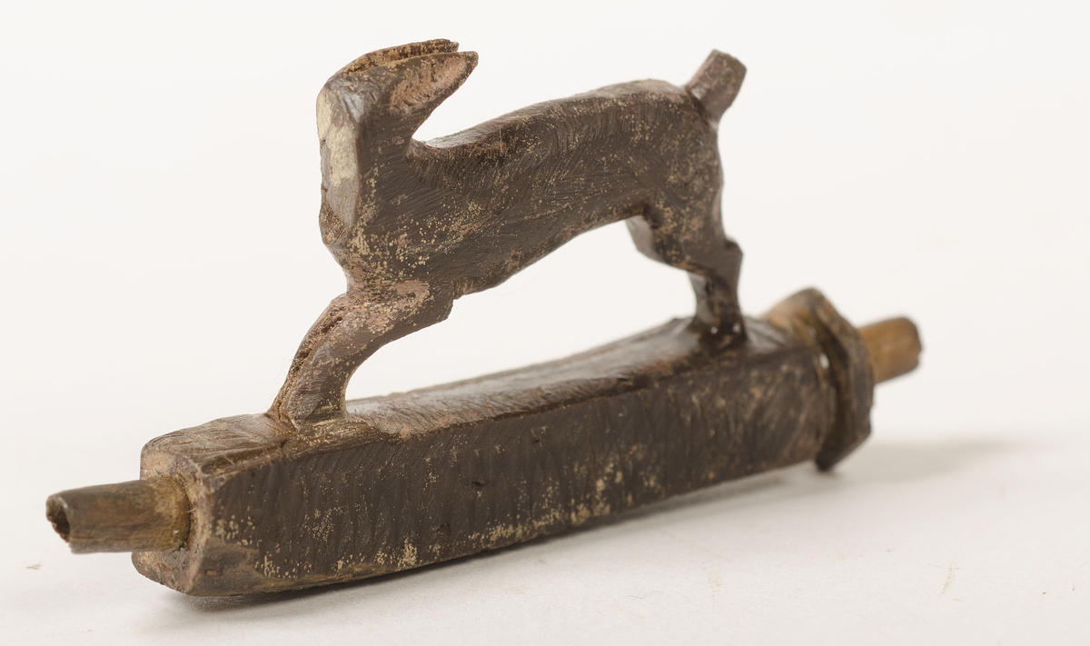 Snidad djurfigur. Basen som djuret står på har kvadratiskt tvärsnitt. Genom basen löper ett ihåligt rör. Djurfiguren föreställer en löpande hare, hjort eller hund. Djurhuvudet är avbrutet.