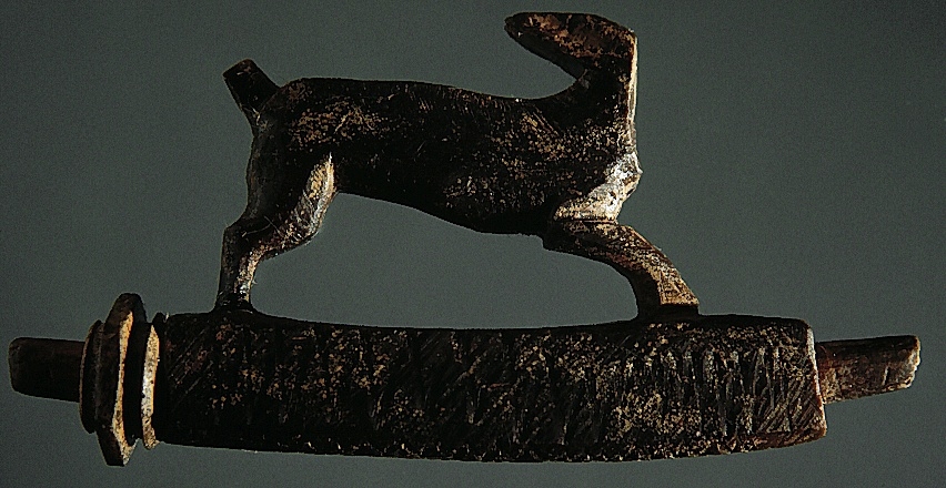 Snidad djurfigur. Basen som djuret står på har kvadratiskt tvärsnitt. Genom basen löper ett ihåligt rör. Djurfiguren föreställer en löpande hare, hjort eller hund. Djurhuvudet är avbrutet.