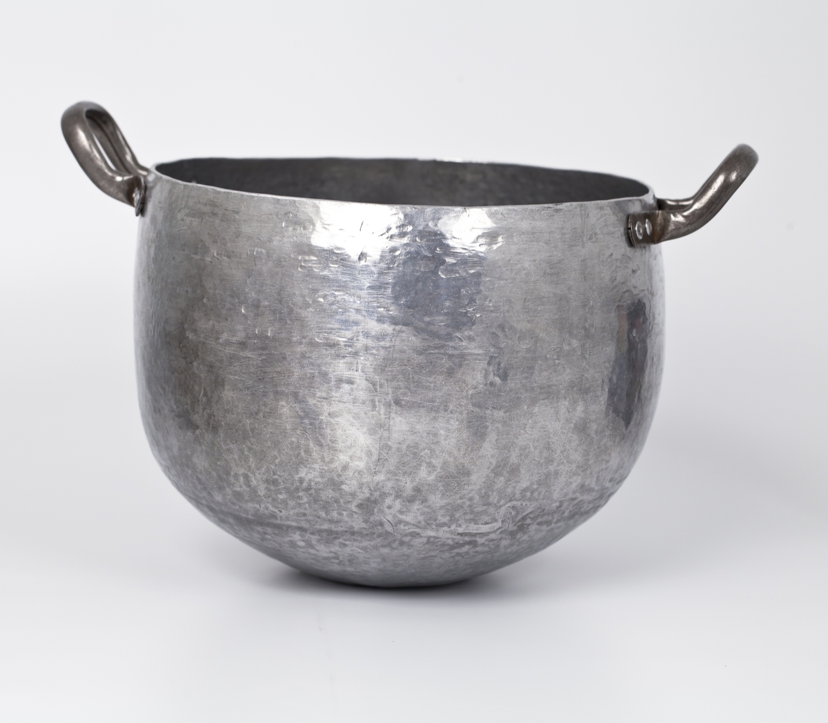 En hamret, høy og oval aluminiumsbolle med et lite ovalt speil i bunnen og to hanker. En korsform i bunnen på undersiden.