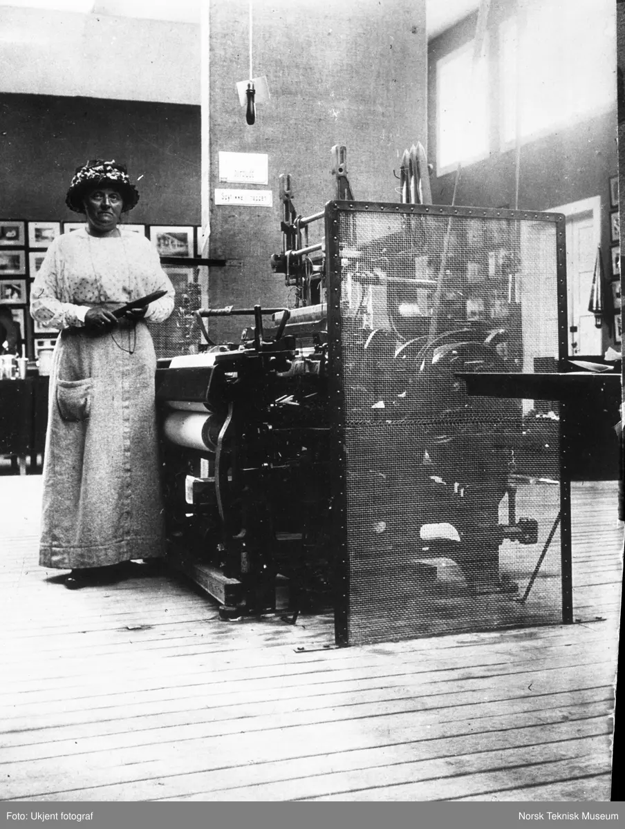 Betzy Kjelsberg ved en vevstol med beskyttelsesgitter, med skyttel i hånden og iført forkle (arbeidstøy). I bakgrunnen skilt der det står "Spyt ikke i trappen". Fra Arbeiderbeskyttelsesutstillingen i Kristiania i 1914.