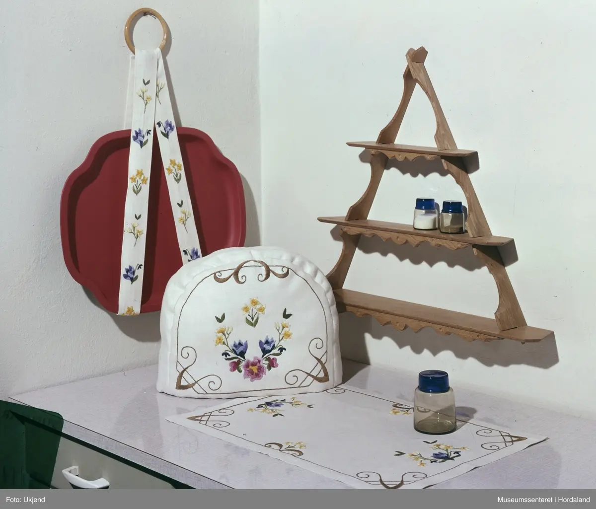 Reklamefoto av brodert løpar, bretthengar og te-hette i interiør, publisert på Spinnesiden i Norsk Ukeblad. Sydd med moulinegarn.