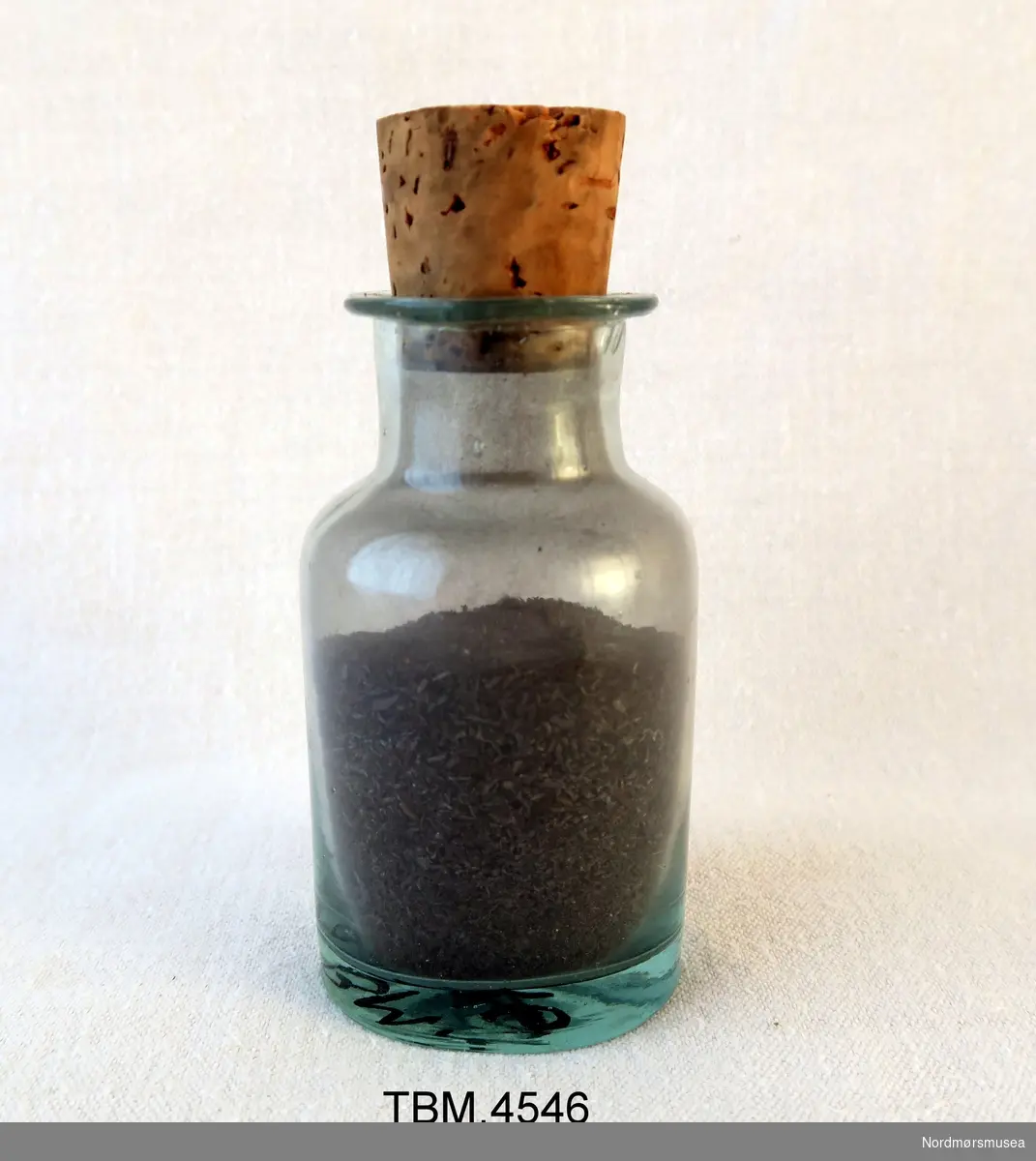 Klar glassflaske med kork. Korken har et hull slik at en kan helle jernfilsponet ut av flasken.
Innhold: Jernfilspon.