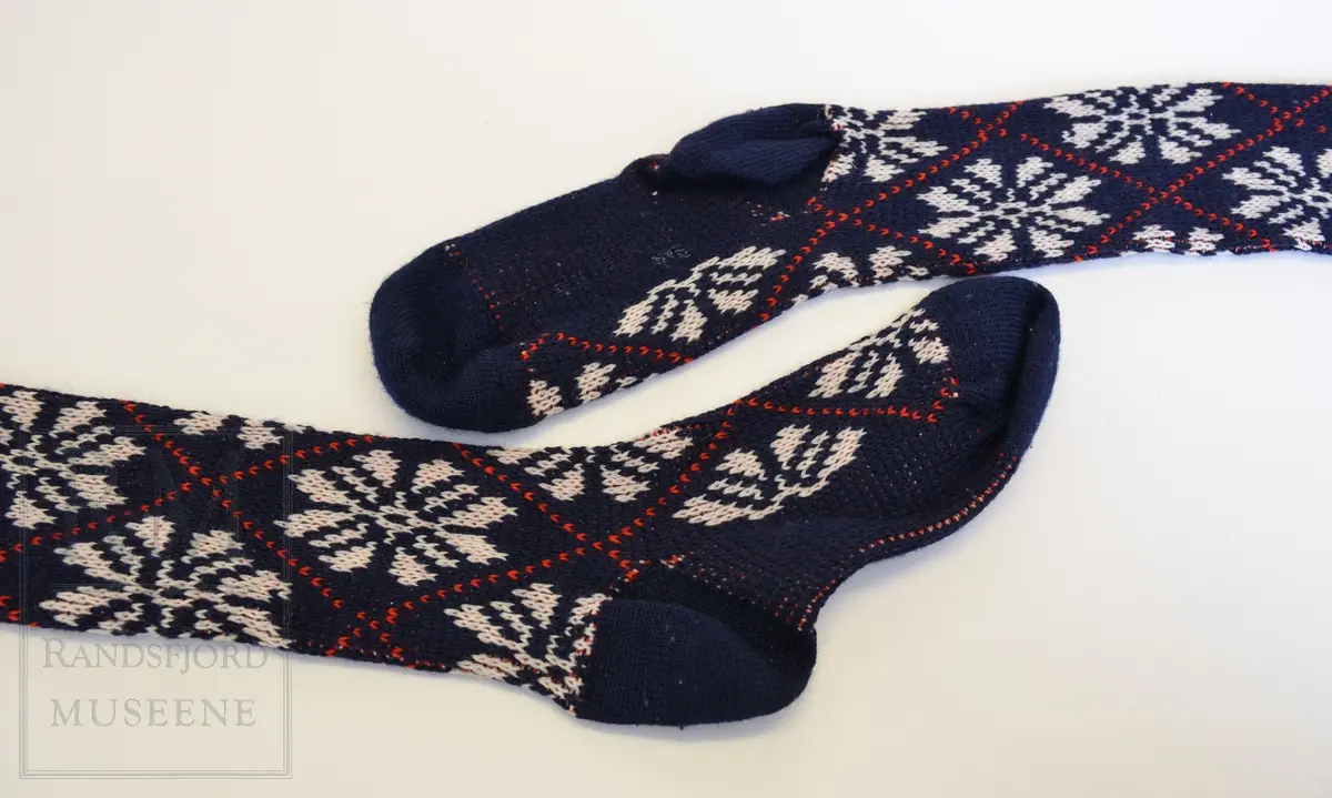 Knestrømper, mønstrede, maskinstrikket, til bruk sammen med knikkers. Herremodell. Ull og kunstfiber. Blå med hvite stjerner/roser og røde striper i rutemønster. Elastikk.