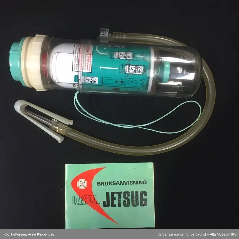 A. Sylinderformet plastapparat med tilfestet gummislange og gjennomsiktig plasttut.  Inneholder en driftspatron fylt med ufarlig gass.
B Rektangulært hefte, bruksanvisning.