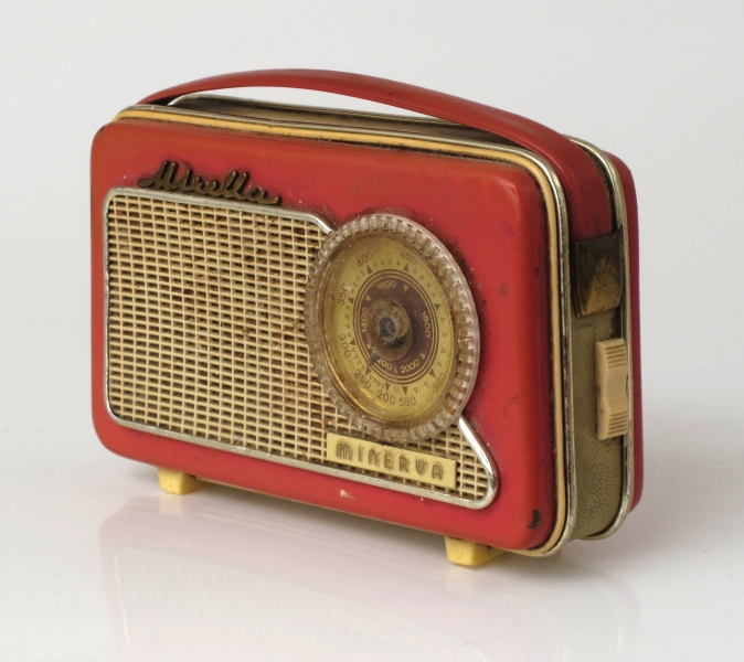 Liten reiseradio i originaleske. Smalt radioapparat  trukket med rosa og elfenbensfarget plast med bærestropp  av rosa plast lærimitasjon. Sirkelrund kanalsøker t. h. på ene langside, med høytalergitter t.v. Ligger i eske av elfenbenshvit kartong med gull , på "pute" trukket m. elfenbens filt.  Med garantikort fra Standard Radiofabrikk.