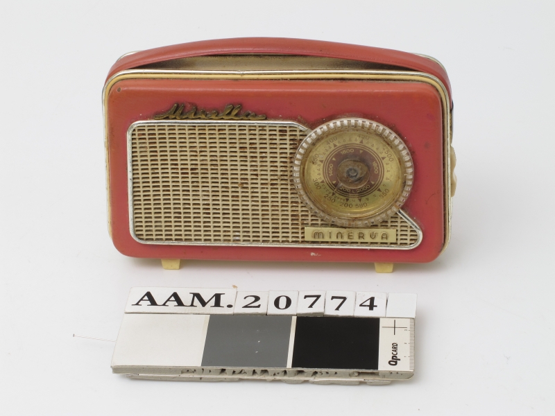 Liten reiseradio i originaleske. Smalt radioapparat  trukket med rosa og elfenbensfarget plast med bærestropp  av rosa plast lærimitasjon. Sirkelrund kanalsøker t. h. på ene langside, med høytalergitter t.v. Ligger i eske av elfenbenshvit kartong med gull , på "pute" trukket m. elfenbens filt.  Med garantikort fra Standard Radiofabrikk.