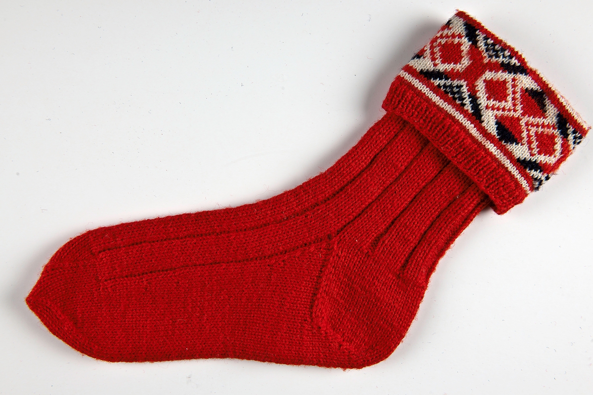 Raggsocka i 4-trådigt rött ullgarn. 1 cm resår sedan blå och röd bjärbobård på vit botten därefter resårstickning 1 r-1a i 3cm sen skaftet 12 cm resår 5 r- 2 a. häl,fot och tå slätstickad