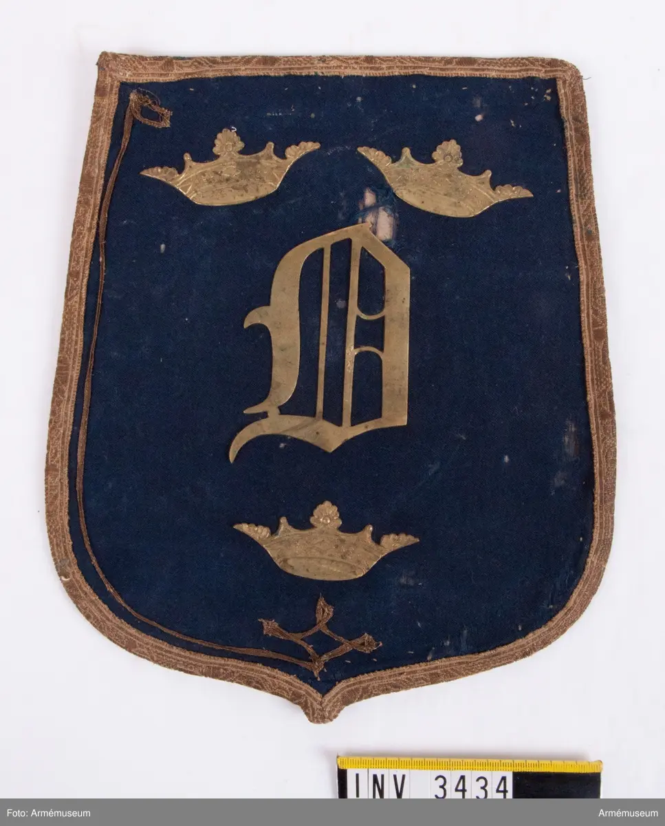 Monogram Oscar I.
Mått: 265 x 220 mm.
Framsidan är täckt  med ett ganska tunt ylletyg, vävt i kypert. Locket är kantat  med ett band i guldgalon. Innanför bandet är som dekoration fastsytt ett mycket smalt guldband, som endast är kvar på ena  sidan och upptill och vid den andra sidan är helt borta. På  mitten av locket är ett 90 mm högt monogram för Oscar I, däröver två öppna kronor och under monogrammet en Öppen krona. Locket är fodrat med ett ljusgult bomullsfoder vävt i tuskaft. Färgen skall väl vara saffianliknande (= marokäng eller saffian, mjukt läder av get eller bockskinn). En ficka finnes på mitten av fodersidan och sträcker sig tvärs  över från sida till sida och går på djupet till lockets nedersta kant. Över ficköppningen fälles ett lock, kantat med  guldband och upptill fastsatt under den guldgalon som går runt  lockets kant. Vilket regemente som haft detta lock med detta utseende vet jag  just nu inte, men gissar på, som hade mörkblå sabeltaska med gul saffian tiden 1814-54. Kanske är det teaterrekvisita? Åke Dalbeck tror "barnleksak" f kungl. barn.