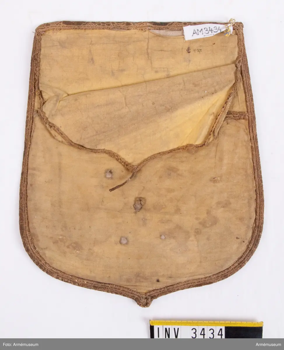 Monogram Oscar I.
Mått: 265 x 220 mm.
Framsidan är täckt  med ett ganska tunt ylletyg, vävt i kypert. Locket är kantat  med ett band i guldgalon. Innanför bandet är som dekoration fastsytt ett mycket smalt guldband, som endast är kvar på ena  sidan och upptill och vid den andra sidan är helt borta. På  mitten av locket är ett 90 mm högt monogram för Oscar I, däröver två öppna kronor och under monogrammet en Öppen krona. Locket är fodrat med ett ljusgult bomullsfoder vävt i tuskaft. Färgen skall väl vara saffianliknande (= marokäng eller saffian, mjukt läder av get eller bockskinn). En ficka finnes på mitten av fodersidan och sträcker sig tvärs  över från sida till sida och går på djupet till lockets nedersta kant. Över ficköppningen fälles ett lock, kantat med  guldband och upptill fastsatt under den guldgalon som går runt  lockets kant. Vilket regemente som haft detta lock med detta utseende vet jag  just nu inte, men gissar på, som hade mörkblå sabeltaska med gul saffian tiden 1814-54. Kanske är det teaterrekvisita? Åke Dalbeck tror "barnleksak" f kungl. barn.