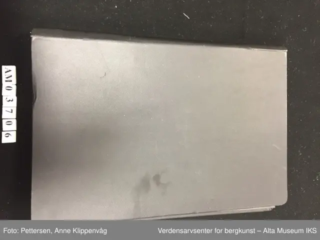 En rektangulær mappe i sort skinn som kan åpnes som en bok. På hver permside er det rammer i metall. I hver ramme er det 33 plastlommer med pappforing. På toppen av hver permside er det en lapp i sort skinn som skjuler den første plastlommen. Under skinnlappen på venstre permsiden er det liten bunke med gule arkivlapper som er håndskrevet på.