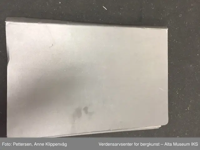 En rektangulær mappe i sort skinn som kan åpnes som en bok. På hver permside er det rammer i metall. I hver ramme er det 33 plastlommer med pappforing. På toppen av hver permside er det en lapp i sort skinn som skjuler den første plastlommen. Under skinnlappen på venstre permsiden er det liten bunke med gule arkivlapper som er håndskrevet på.
