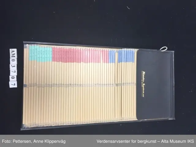 En rektangulær sort ramme i metall som er stiftet på et styrkke papp trukket i sort skinn. I rammen er det 49 plastlommer med pappforing. På toppen er det en lapp i sort skinn som skjuler den første plastlommen. På venstre siden av hver enkel plastlomme er det en liten farget lapp med påskrift.