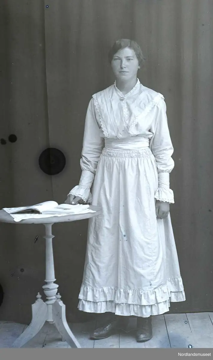 Portrett. Jente i tenårene. Står ved et lite bord. Samme jente som på NM 8243:585.