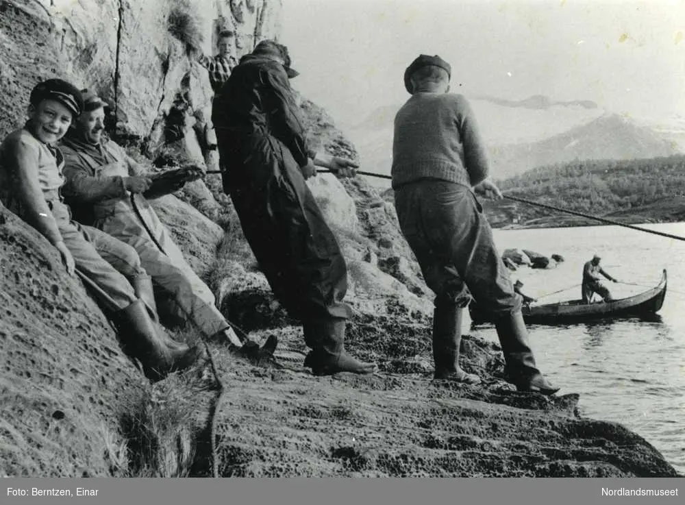 Fiske med landnot under Bratthammaren ved kongestøtta. Notlaget til Egil Olsen, Bratthammar. En straumbåt med to personer ombord som trekker i et tau. På land står tre voksne menn og trekker i et tau. En gutt står og ser på. Vannet står nesten stille.