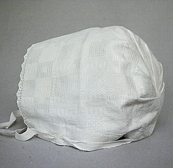 Huvudbonad sk käringhatt till Leksandsdräkt. Sydd i mönstervävt linne i daldrällsbindning, kantad framtill med virkad spets i bomull. Broderat blommönster mitt bak vid veckningen. Vitt bomullsband som löper genom kanal och langetterade hål.