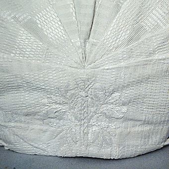 Huvudbonad sk käringhatt till Leksandsdräkt. Sydd i mönstervävt linne i daldrällsbindning, kantad framtill med virkad spets i bomull. Broderat blommönster mitt bak vid veckningen. Vitt bomullsband som löper genom kanal och langetterade hål.