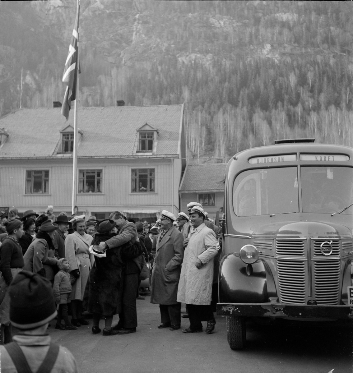 Bjørnson-koret holder konsert på Rjukan torg. Medlemmer av koret med hvit lue stående på utsiden av buss merket med Bjørnson-koret. Mennesker stående rundt bussen.