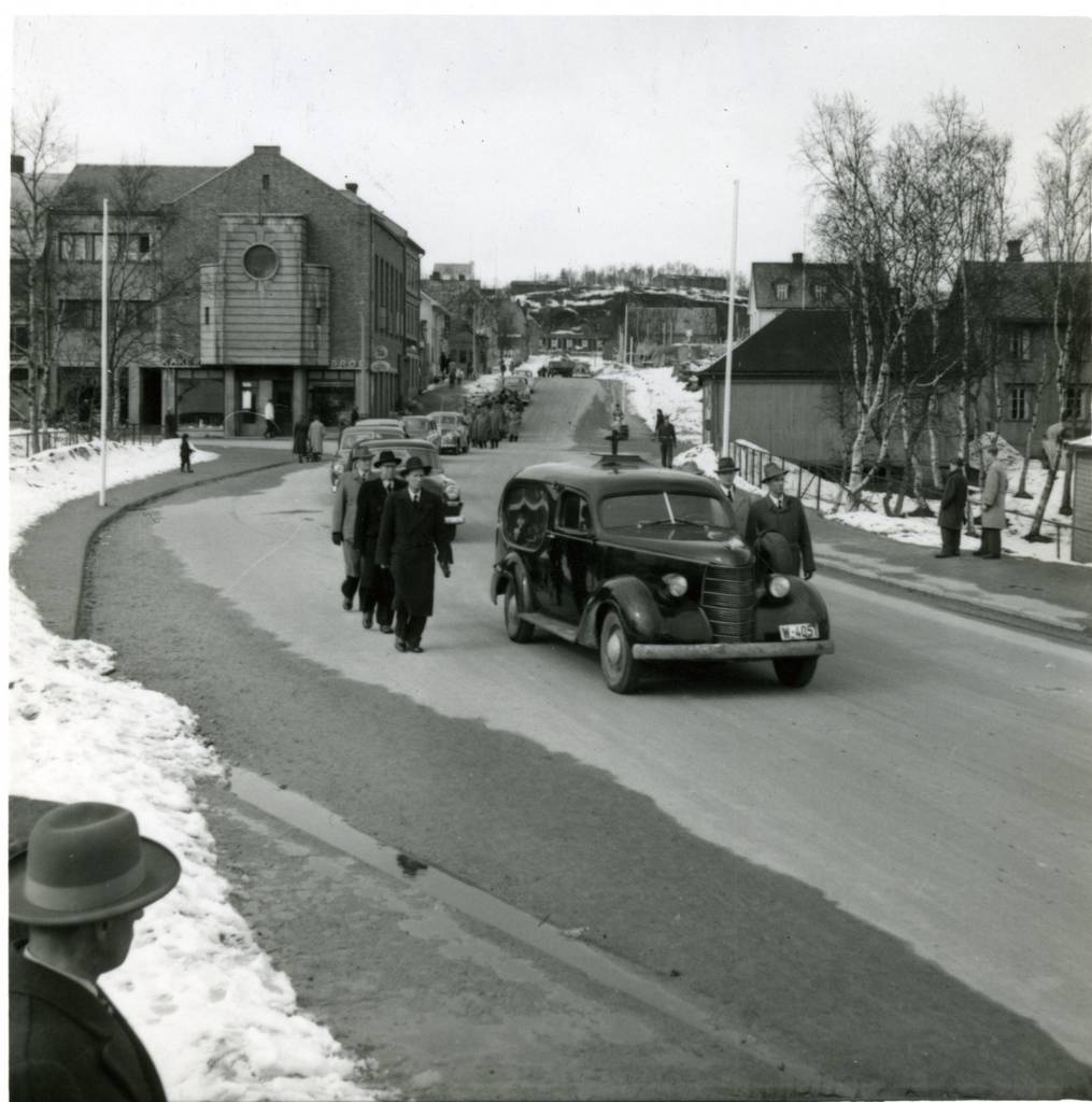 Narvik, Begravelsesfølge fra Narvik Kirke. I Brugata krysset med Frydenlundsgata. Bærere og gravfølget fulgte kisten til Narvik gravlund i Taraldsvik. Begravelsesbilen tilhørte sansynligvis Frydendal.  Til venstre Baptistkirken,og bak den ser vi slakter Lians gård. Til høyre Levin (Skotøykristiansen) og bak den og oppe på "Tjyvberget" ser vi den store hvite gården til Langsether. Madam Johnsen-stua er revet.  Bakerst i Brugata er det satt opp tyskbrakke.
Bildet må i alle fall være tatt etter mai 1947, da Hvem eier bilen 1947 var ajour. Der står W-4051 fortsatt på en Chevrolet lastebil som tilhørte Åsmund Åsfjord, Leines i Steigen. Og enda nyere: Bilhistoriker Asbjørn Rolseth påpeker at bil nr 2 med hel frontrute må vel være nyere enn 1950, det lille vi ser tyder på en 1953 Plymouth (eller Dodge Kingsway)?