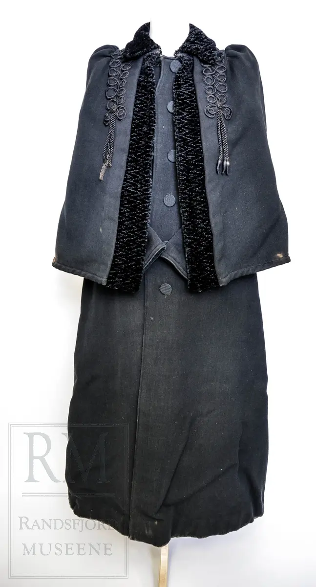 Sort kåpe i ullstoff med påsydd cape. Venstrekneppet, 6 stofftrukne knapper. Dekorert med flettede bånd (agraman). Lang, innsvinget modell med splitt bak. Uforet.