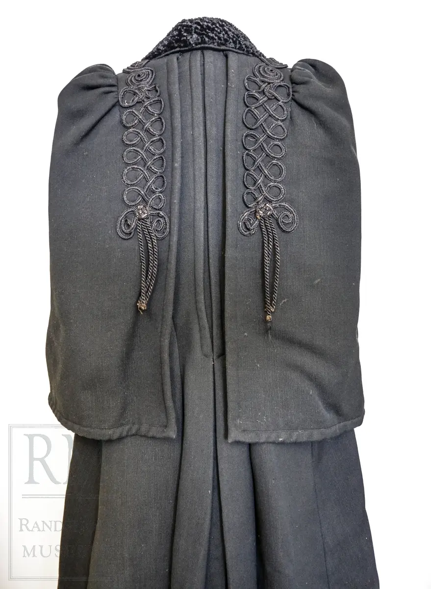 Sort kåpe i ullstoff med påsydd cape. Venstrekneppet, 6 stofftrukne knapper. Dekorert med flettede bånd (agraman). Lang, innsvinget modell med splitt bak. Uforet.