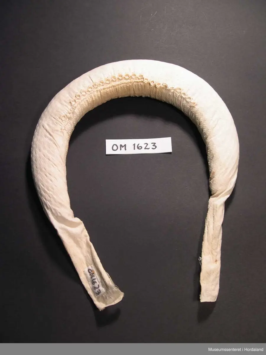 Boga og runda valk i U-fasong med fyll, trekt med kvitt bomullsstoff. Fyllet går ikkje heilt ut til endane. Skal setjast oppå hovudet og brukast under kvithua for å få rett fasong og for å løfta hua vekk frå ansiktet. Festast i nakken. Påsydd ei lita bord av nupereller midt på. Denne skal koma midt oppå hovudet framme.