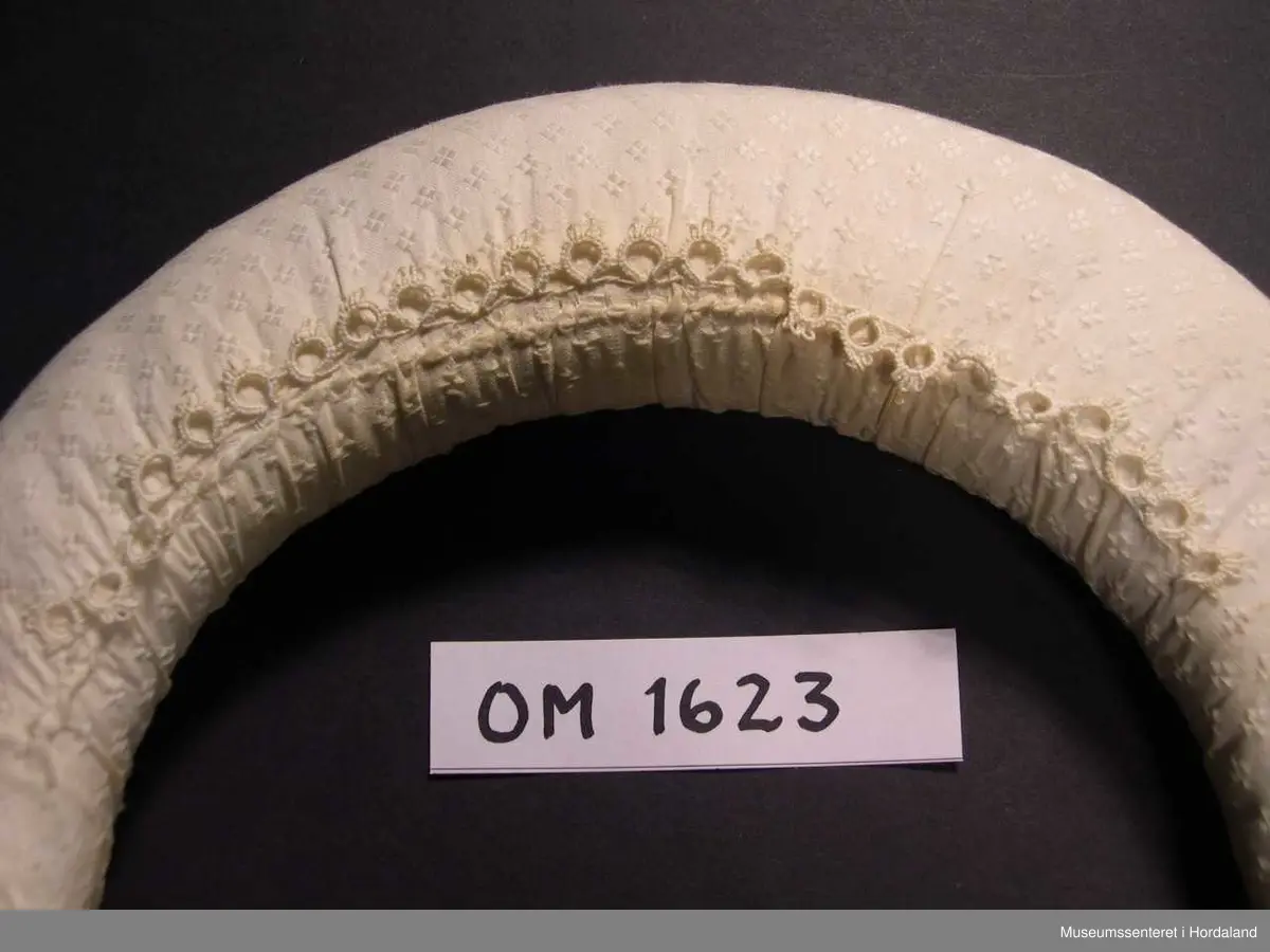 Boga og runda valk i U-fasong med fyll, trekt med kvitt bomullsstoff. Fyllet går ikkje heilt ut til endane. Skal setjast oppå hovudet og brukast under kvithua for å få rett fasong og for å løfta hua vekk frå ansiktet. Festast i nakken. Påsydd ei lita bord av nupereller midt på. Denne skal koma midt oppå hovudet framme.