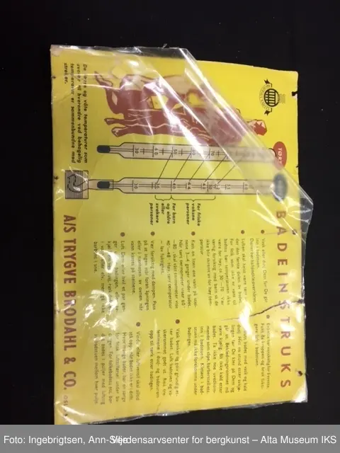 En rektangulær plakat i gul papp. Skrevet på med rød og svart skrift på høyre side. På venstre side er det en tegning med instruksjoner. Innpakket i plast.