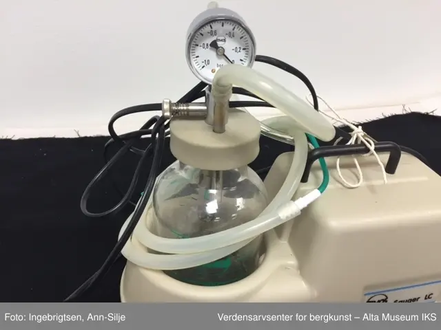 Rektangulært apparat med avrundet sokkel med en sylinderformet glasskolbe i. På lokket til glasskolben er det en trykkmåler og to metalltuter hvor det er festet en gummi/plastslange i hver. Den ene slangen går inn i selve apparatet og den andre er kveilet rundt glasskolben. Glasskolben er merket med grønne streker og tall. På venstre siden av apparatet er det en av- og på-knapp. Bruksområdet er å suge opp væske, særlig i fra luftveiene.