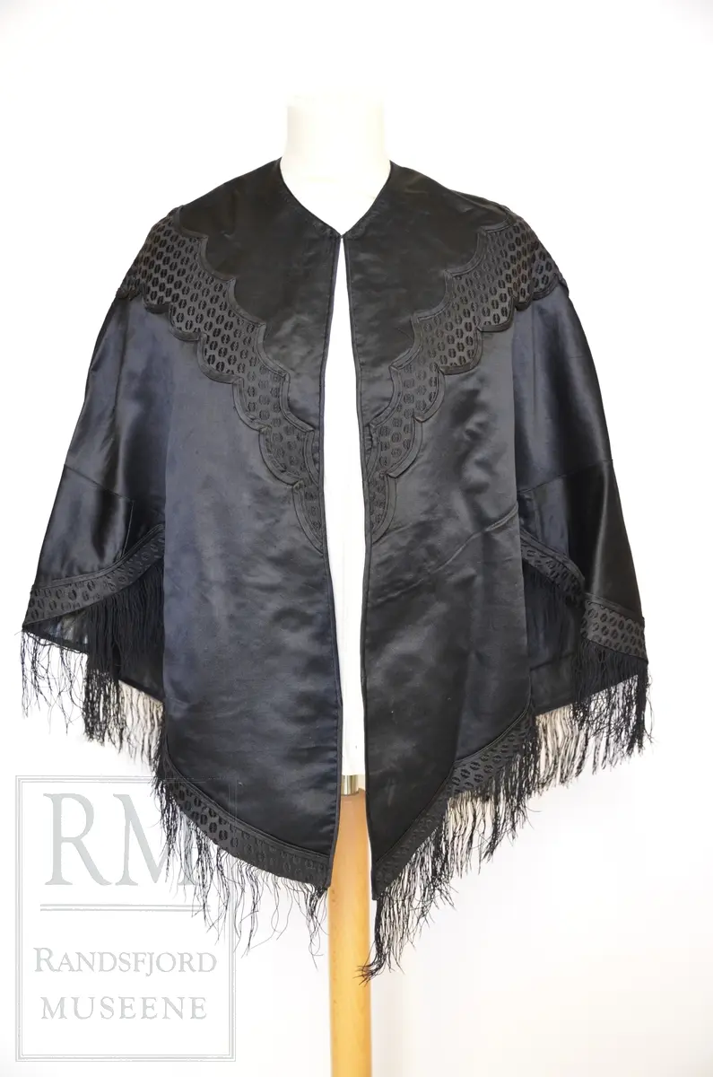 Håndsydd. Svart silkesatin med glanset bakside. Alpakkarenning (?) m / innslag i natursilke. Applikasjon av svart mønstret silkerips. To agramanrosetter med svarte glassperler. Sammensydd i rygg. Lukkes med hekte og malje ved halsåpning foran. Frynser.