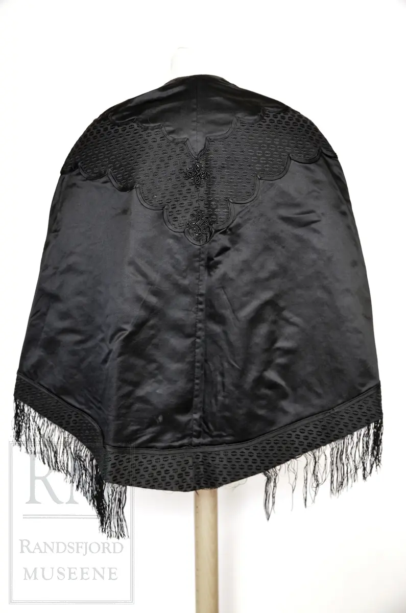 Håndsydd. Svart silkesatin med glanset bakside. Alpakkarenning (?) m / innslag i natursilke. Applikasjon av svart mønstret silkerips. To agramanrosetter med svarte glassperler. Sammensydd i rygg. Lukkes med hekte og malje ved halsåpning foran. Frynser.