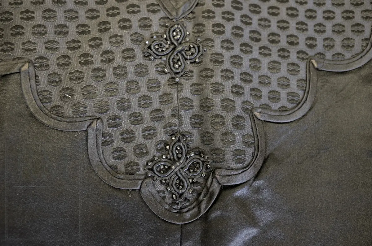 Håndsydd. Svart silkesatin med glanset bakside. Alpakkarenning (?) m / innslag i natursilke. Applikasjon av svart mønstret silkerips. To agramanrosetter med svarte glassperler. Sammensydd i rygg. Lukkes med hekte og malje ved halsåpning foran. Frynser.