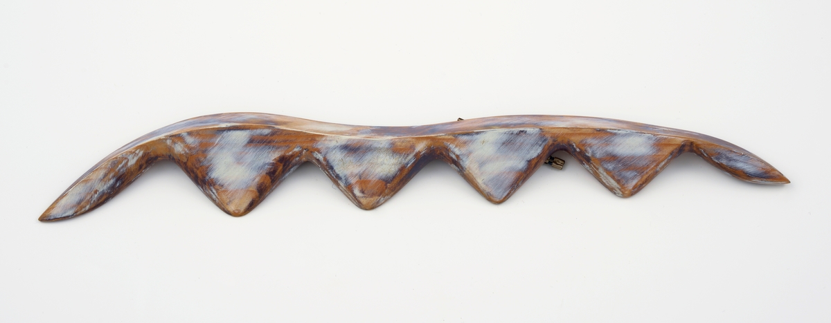 Avlang brosje i balsatre med trekantet tverrsnitt. Oversiden har bølgeform langs den ene kanten, sagtannsnitt langs den andre. Oversiden er flekkvis malt med hvit og blålig maling. Brosjen har to stålklips på baksiden.