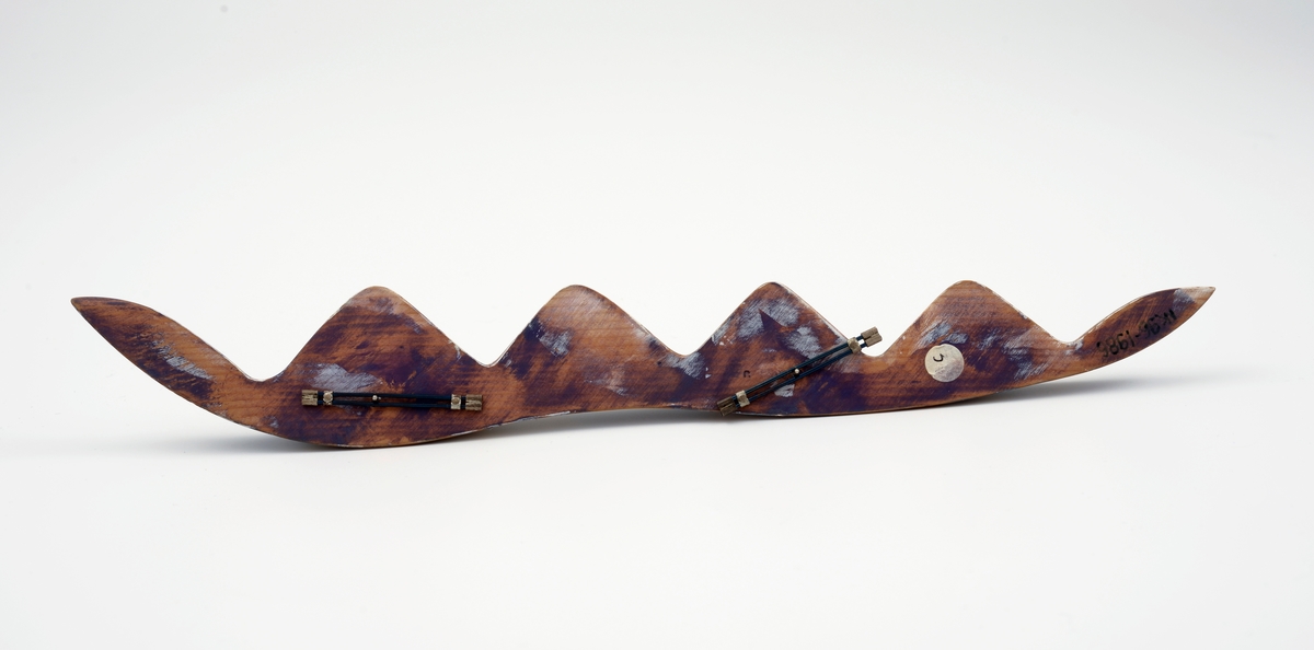 Avlang brosje i balsatre med trekantet tverrsnitt. Oversiden har bølgeform langs den ene kanten, sagtannsnitt langs den andre. Oversiden er flekkvis malt med hvit og blålig maling. Brosjen har to stålklips på baksiden.