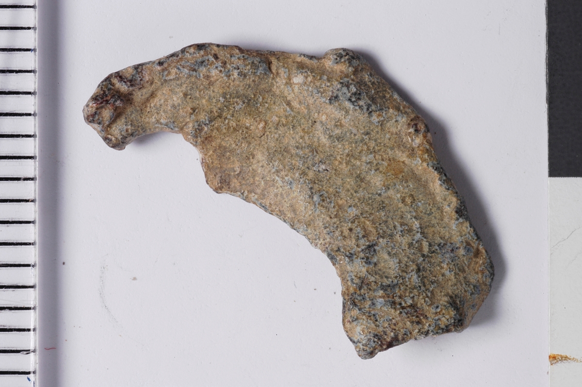 Del av plakett fra vareplombe, som har hatt to nagler. Gjenstanden har tegn til preg i form av perlerand i ytterkant. 