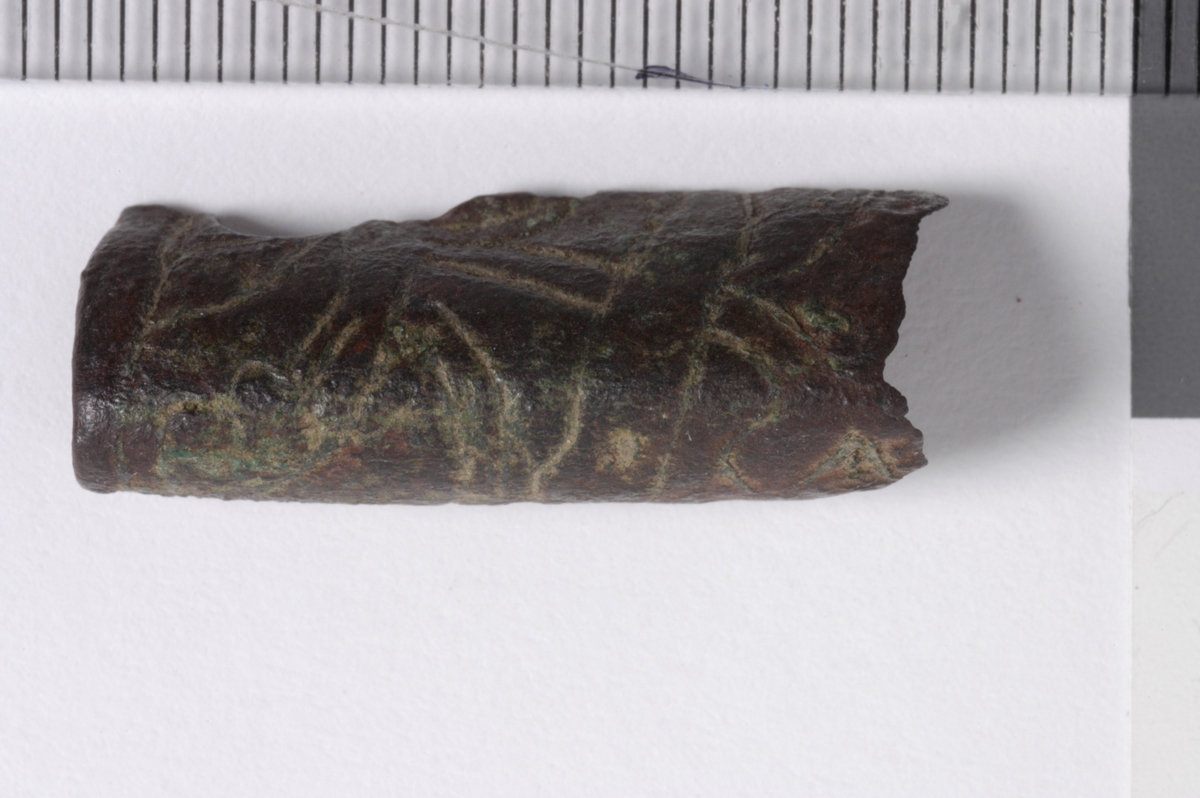 MALV: L. 25 MM, BÅNDFORMET FRAGMENT SOM ER BØYET. OVERSIDEN HARINNPRESSET DEKOR. DEN ENE ENDEN BEARBEIDET. 