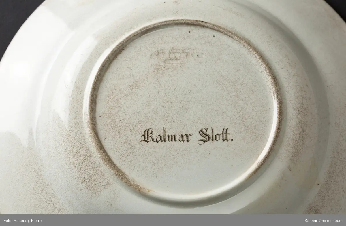 KLM 45642 Tallrik, av porslin. Djup tallrik med tämligen brett brätte. På undersidan text: Kalmar slott, samt tillverkarstämpel som inte kunnat tydas. Tryckt dekor med motiv av Kalmar slott i brunt och grönt, slottet inbäddat i grönska, nedanför vallarna buskar och träd. Motivet dateras till 1860-70-tal, eftersom tornhuven på Kuretornet omgestaltades ca 1880. Om motivet och tallriken är från samma tid, kan tallriken dateras till 1860-70-tal.