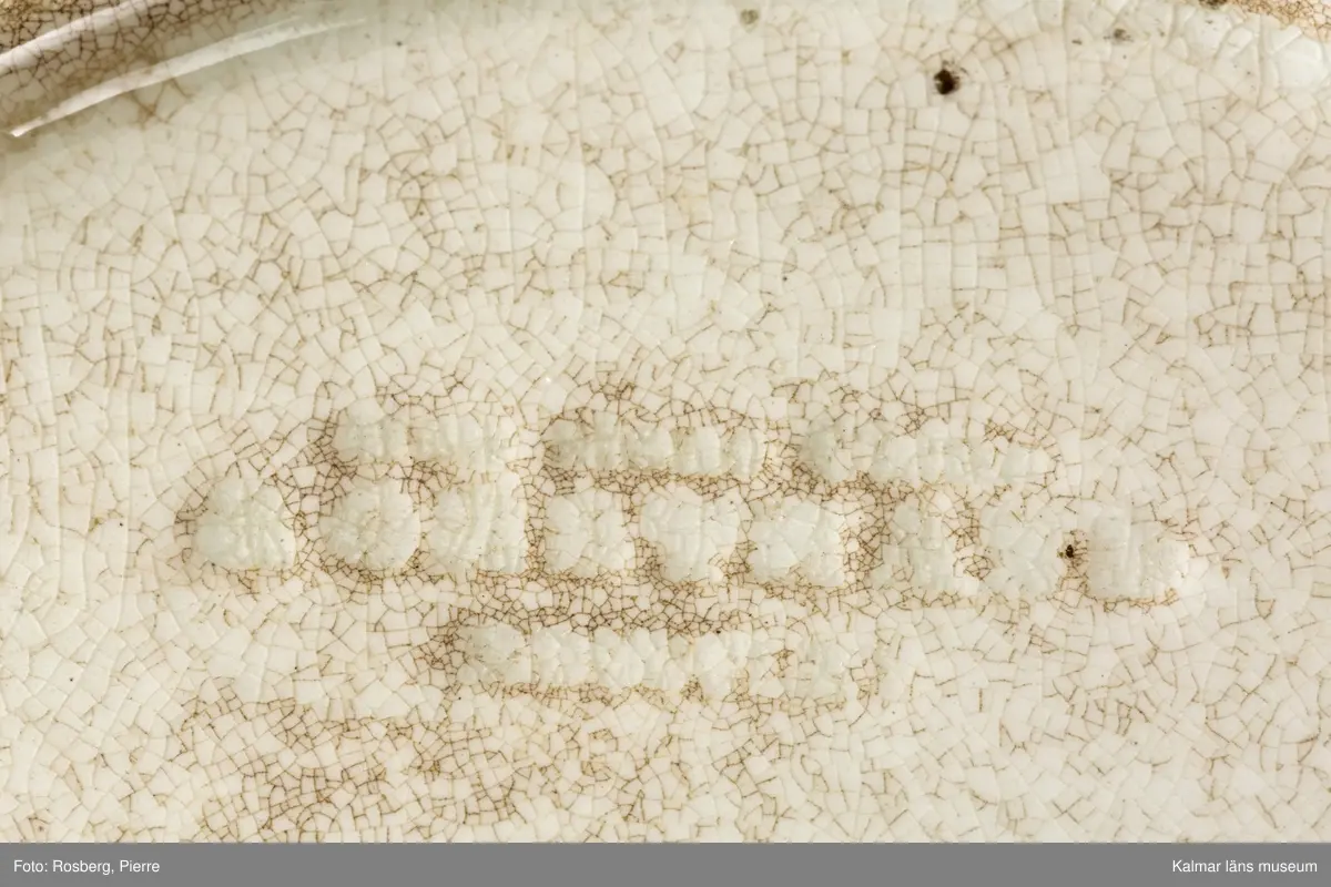 KLM 45642 Tallrik, av porslin. Djup tallrik med tämligen brett brätte. På undersidan text: Kalmar slott, samt tillverkarstämpel som inte kunnat tydas. Tryckt dekor med motiv av Kalmar slott i brunt och grönt, slottet inbäddat i grönska, nedanför vallarna buskar och träd. Motivet dateras till 1860-70-tal, eftersom tornhuven på Kuretornet omgestaltades ca 1880. Om motivet och tallriken är från samma tid, kan tallriken dateras till 1860-70-tal.