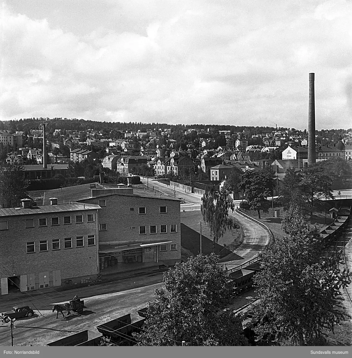 Stadsvy fotograferad från hamnen, Järnvägsparken, mot Södermalm. Närmast syns korsningen Köpmangatan/Landsvägsallén och Parkgatan samt järnvägen som går under Köpmangatan vid gasverket. I nedre vänstra hörnet syns en man som kör en häst ner mot hamnen.