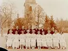 Konfirmander, 10 flickor, 7 pojkar och kyrkoherde Jäderlund.
Rinkaby kyrka i bakgrunden.